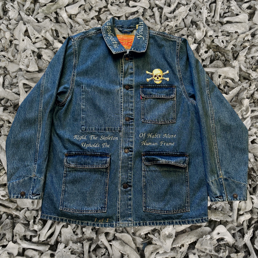Unapologetic Youth denim jean jacket with bone graphic design, classic fit, and button-front closure, displayed on a hanger.